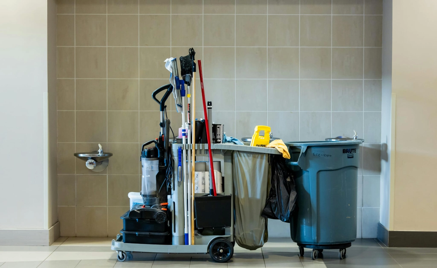 R&O Facilityservice – Ein Reinigungswagen, beladen mit Mopps, Besen, Reinigungsmitteln und einem Mülleimer, steht in einem gefliesten Flur neben einem Trinkbrunnen.