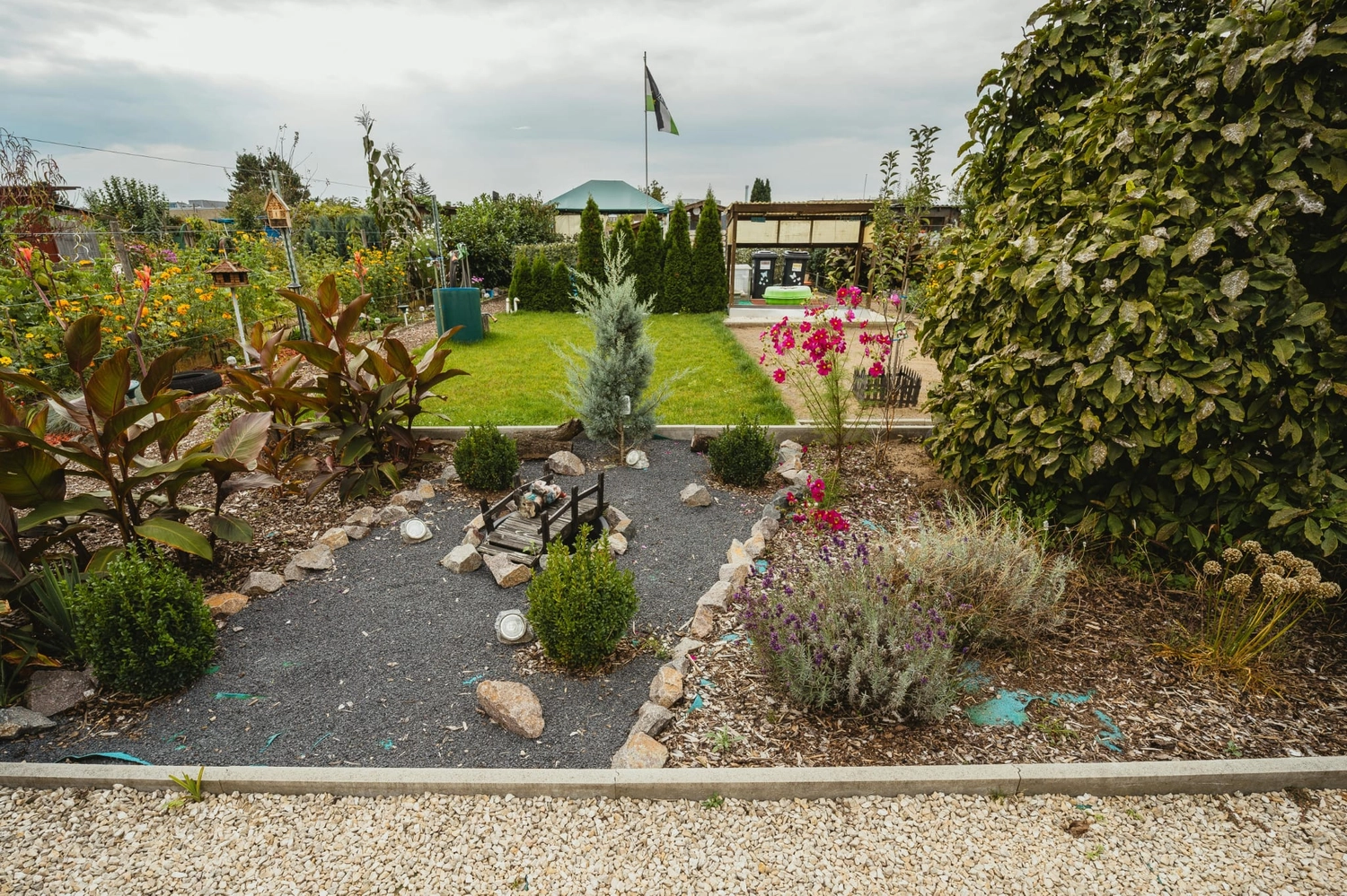 R&O Facilityservice – Ein gepflegter Garten mit Kieswegen, Sträuchern, bunten Blumen und einem grünen Rasen unter bewölktem Himmel.