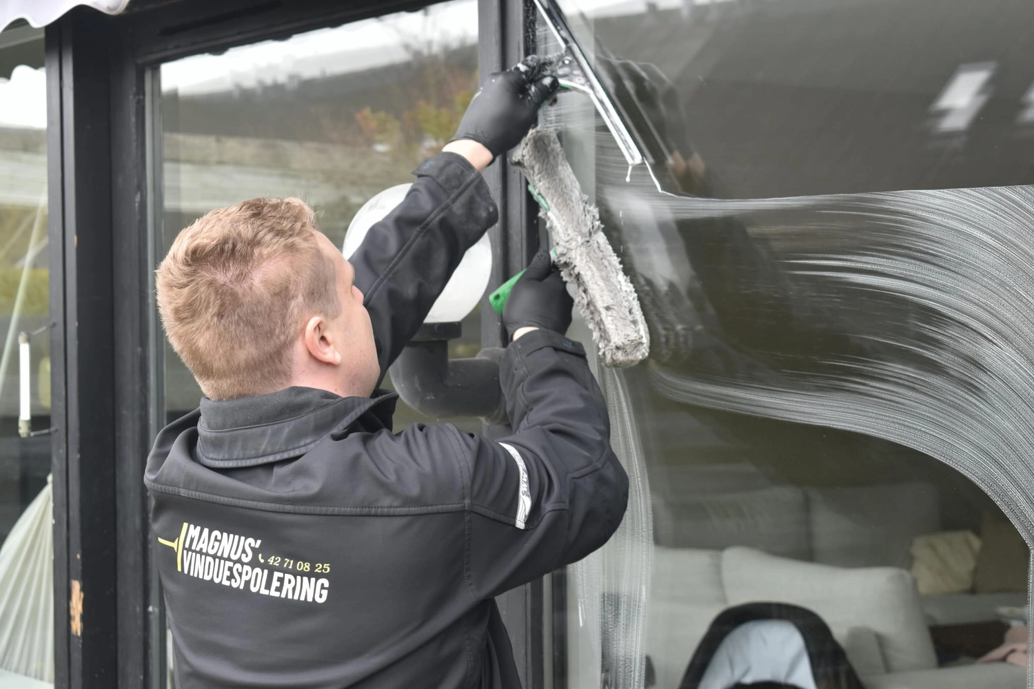 R&O Facilityservice – Ein Fensterputzer in schwarzer Uniform, der mit einem Seifenreiniger ein großes Glasfenster schrubbt.